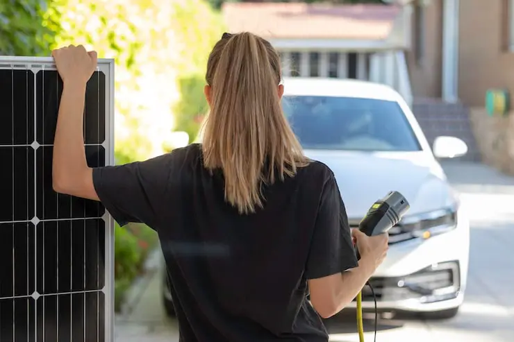 A woman holds an EV charging plug in one hand standing in front of a white EV.