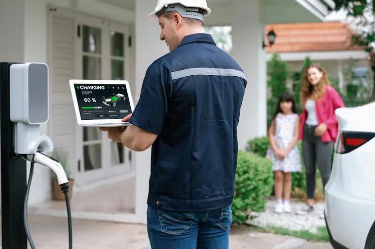 An electrician in a hard hat and uniform is checking the charging status on a laptop near an EV charger and a white electric car.