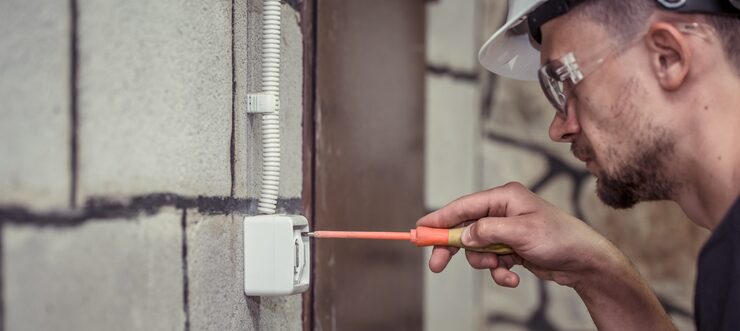 Protect Home From Electrical Surges in Ontario (4) An electrician wearing a hard hat and safety goggles is checking an electrical outlet with a screwdriver to perform troubleshooting of faulty wiring that can lead to power surges.