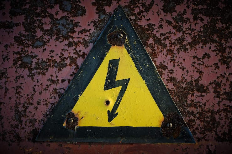 Signs Electrical Panel Damaged in a Storm (1) A close-up of a weather-beaten, yellow and black triangular electrical hazard sign with a lightning bolt, bolted onto a heavily rusted red-brown metal surface, suggesting damage and decay, possibly due to a storm.