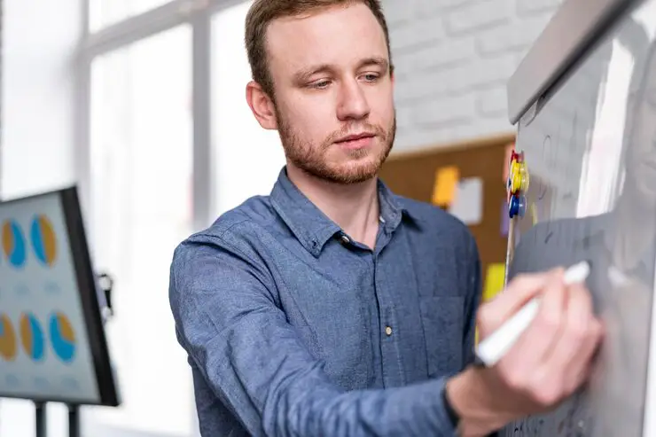 A focused, clean-shaven man in a blue button-up shirt is writing with a marker on a white flip chart.