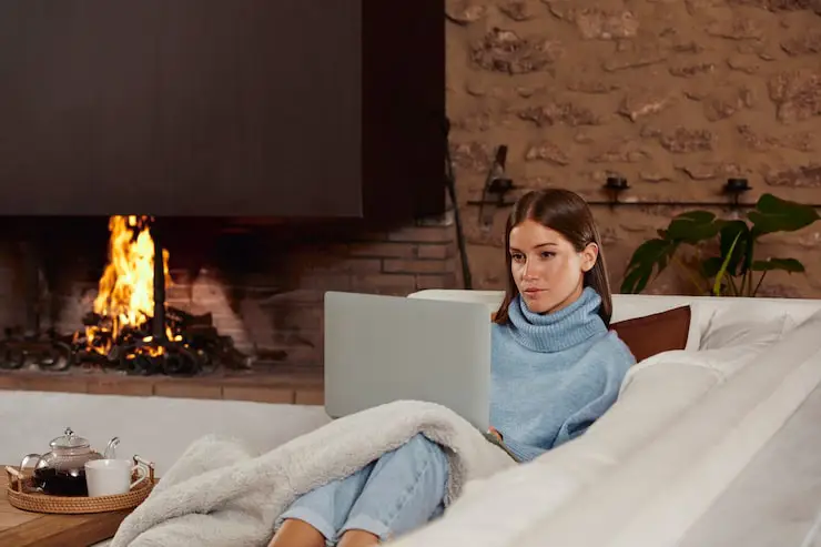 Woman cozy on a couch with a laptop and a blanket next to a fireplace.
