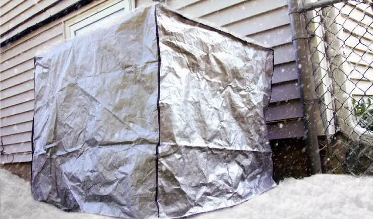 Silver AC cover protecting unit from falling snow against house siding.